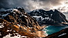Snødekte fjell over en turkis fjord med smal strand og dramatiske skyer.