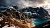 Snødekte fjell over en turkis fjord med smal strand og dramatiske skyer.