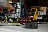 En liten svart boks står på et bord blant kabler og testutstyr, mens en hånd arbeider ved elektronikk i bakgrunnen.